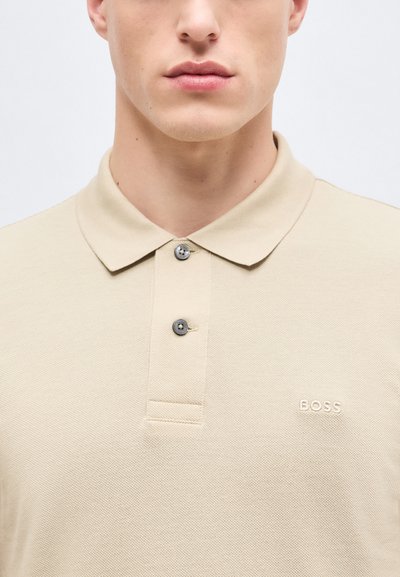 Jeune homme portant un polo beige BOSS avec deux boutons fermés, visible du cou jusqu'au milieu de la poitrine, sur un fond uni.