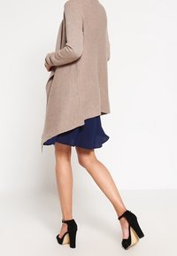 Cardigan beige a coste con un orlo asimmetrico, indossato sopra un abito svasato blu scuro. Scarpe con tacco alto nere completano l'outfit.