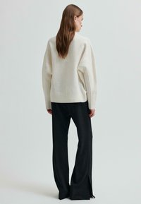 Kobieta o długich brązowych włosach, ubrana w luźny biały sweter i szerokie czarne spodnie, stoi plecami do widza na jednolitym jasnym tle.