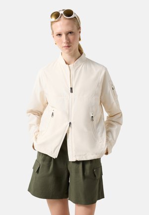 Blondynka w kremowej kurtce na zamek, oliwkowo-zielonych szortach bojówkach i okrągłych okularach przeciwsłonecznych spoczywających na głowie, z rękami w kieszeniach.