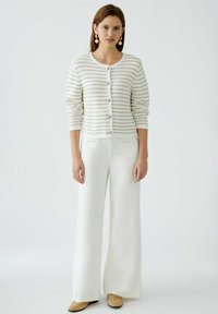 Button-up cardigan in white and light beige stripes, made of knit material. Paired with wide-leg white pants and beige shoes.