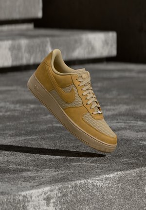 Ruda ir smėlio spalvos Nike Air Force 1 sportbačiai, sklendžiantys virš betono paviršiaus su akmeninėmis laipteliais fone.