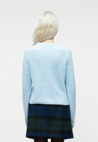 Oseba z dolgimi blond lasmi, oblečena v svetlo modro pulover in temno modro-zeleno karirano krilo, prikazana s hrbta na bež ozadju.