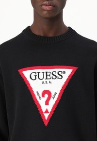 Juodas megztinis su dideliu raudonu ir baltu trikampio emblema su užrašu "GUESS" ir "?". Tekstūruota medžiaga su raukšlėtu apykaklės kraštu.