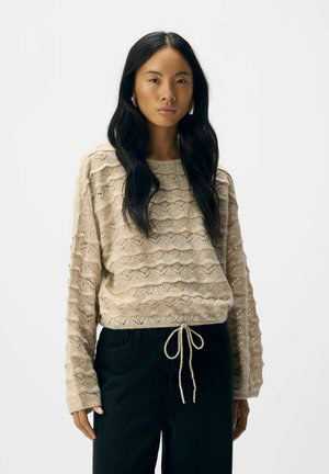 Frau mit langen schwarzen Haaren, die einen beigen strukturierten Strickpullover mit Kordelzug und schwarze Hosen trägt, steht vor einem schlichten weißen Hintergrund.
