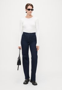 Jeans escuros de cintura alta com perna reta, combinados com uma blusa justa de manga longa branca. Os acessórios incluem uma mala preta e óculos de sol.