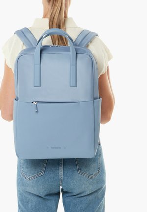 Person wearing light blue square-shaped backpack with top handles and zipped front pocket, dressed in white shirt and blue jeans.