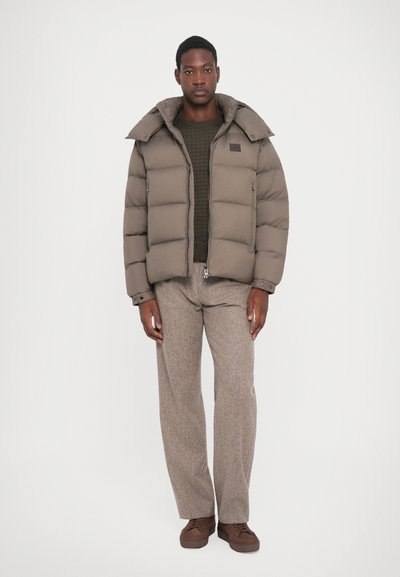 Puffer jacket in olive green with a high collar, textured fabric, and pockets. Paired with brown patterned trousers and matching shoes.