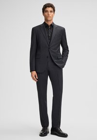 Donkergrijs op maat gemaakt pak met een notch-lapel, twee voorzakken, slim-fit broek, gecombineerd met een zwart overhemd met knopen en zwarte dresschoenen.