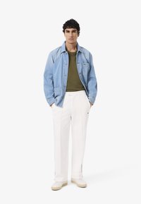 Hombre con chaqueta de mezclilla azul claro, suéter verde olivo, pantalones blancos y zapatillas blancas, de pie con las manos en los bolsillos.