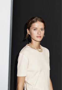 Beige bomulds t-shirt med korte ærmer, prydet med en tekstureret halskæde lavet af runde træperler. Baggrund: mørkegrå.