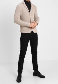 Beige stickad blazer med ett strukturerat mönster, enstaka knappstängning, två framfickor, kombinerad med svarta tajta byxor och svarta stövlar.