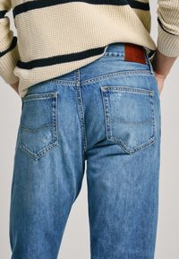 Vaaleansiniset farkut, joissa on kulutettu viimeistely, ruskea nahkapaikka vyötäröllä ja kaksi takataskua, joissa on ommeltu yksityiskohta.