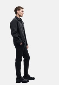 Schwarzes, strukturiertes, knopfbares Hemd, kombiniert mit schwarzen, schmal geschnittenen Hosen und schwarzen Schnürschuhen. Das Modell steht im Profil vor einem weißen Hintergrund.