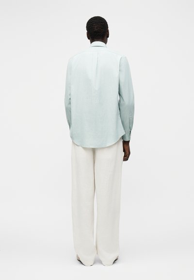 Person med kort krøllete hår sett bakfra, iført en lyseblå langermet skjorte og løse hvite bukser mot ensfarget bakgrunn.