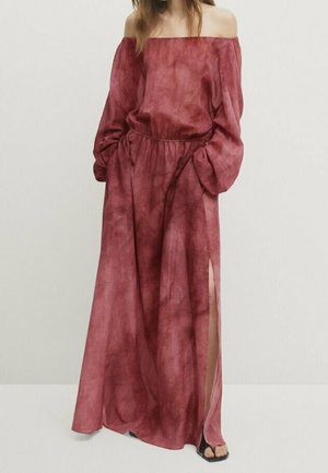 Femme portant une longue robe bordeaux, épaules dénudées, manches longues et une fente haute sur le côté, debout les mains dans les poches.
