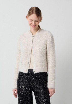 Light pink textured cardigan with a round neckline and silver buttons, featuring a wavy pattern, paired with black sequin pants.
