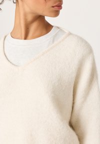 Krémový svetr s výstřihem do V a měkkou, načechranou texturou, s širokými rukávy a neupraveným okrajem. Nasažený přes světle šedou košili.