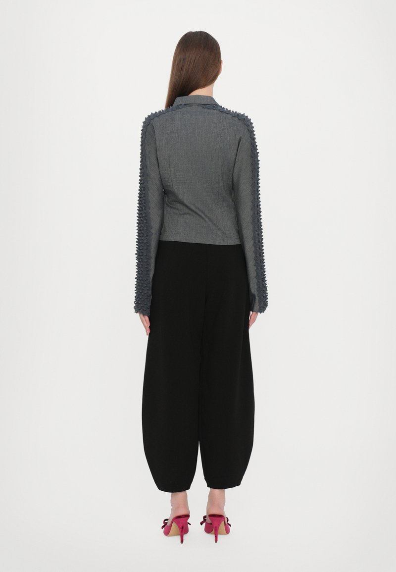 Grå kort jacka med texturerade, spikiga ärmar, i kombination med vida svarta byxor och rosa skor med rosetter. Bakifrån.