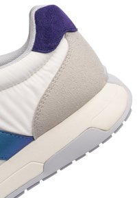 Gros plan sur le talon d'une basket en daim gris, avec un rembourrage blanc, une languette en daim violet, un accent bleu et une semelle en caoutchouc grise.