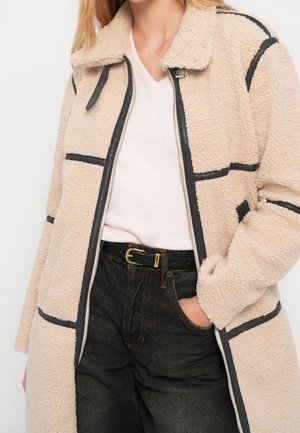 Veste sherpa beige avec bordure noire sur un top blanc, associée à une ceinture noire et un jean en denim foncé, main dans la poche.