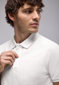 Bílý polo tričko s texturovanou tkaninou, na levé straně s malým logem a tradičním límcem. Model drží límec.