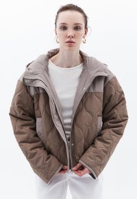 Prošívaná hnědá péřová bunda s kapucí, zipem a knoflíkovým uzávěrem, která má hladké, kontrastní světle šedé rukávy a texturovaný povrch.