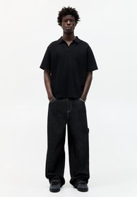 Polo nero a maniche corte abbinato a pantaloni in denim nero larghi. L'outfit presenta un design minimalista e dettagli di cucitura sottili.