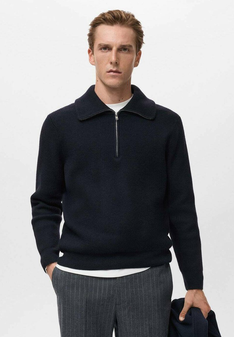 Pullover lavorato a maglia navy con colletto alto e chiusura a zip a quarto. Tessuto a coste, maniche fitted, indossato sopra una camicia bianca. Pantaloni a righe verticali.