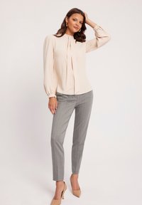 Bladoróżowa bluzka z długimi, bufiastymi rękawami i wiązaniem z przodu, połączona z szarymi spodniami w prążki i cielistymi szpilkami.