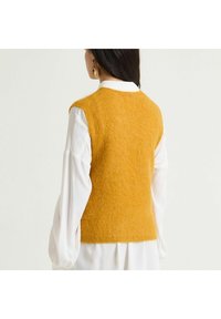 Pull sans manches en tricot jaune moutarde avec tissu texturé, col rond et coupe décontractée, superposé sur une chemise blanche à manches longues.