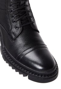 Botín de cuero negro con superficie texturizada, puntera lisa y suela robusta con diseño resistente. Los cordones se extienden hasta el tobillo.
