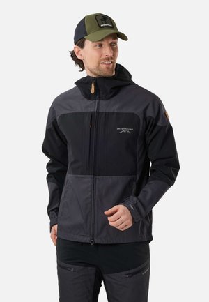 Mann trägt eine schwarze und dunkelgraue Swedemount Outdoorjacke und Hose sowie eine olivgrüne und schwarze Kappe, schaut zur Seite.