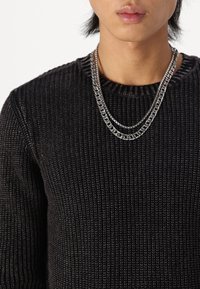 Jeune personne portant des colliers en argent superposés et un pull en maille noire texturée, montrée des épaules jusqu'aux lèvres sur un fond blanc.