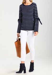 Námořnický pruhovaný svetr s dlouhými rukávy a na zavázání, doplněný bílými slim-fit kalhotami a tmavě modrými kotníkovými botami, držící béžovou kabelku.