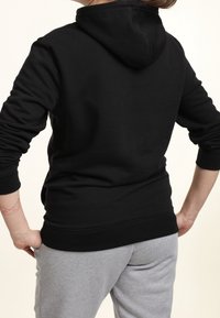 Sweat à capuche noir en tissu doux avec des poignets et un ourlet côtelés. Porté avec un pantalon de jogging gris clair. La vue arrière montre la capuche et les coutures.