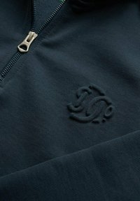 Sweat à zip bleu marine en tissu texturé avec un logo embossé discret sur la poitrine gauche et une fermeture éclair en métal.