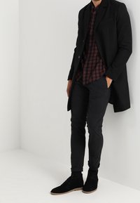 Zwarte jas, rode en zwarte geblokte overhemd, slanke zwarte broek en zwarte suède laarzen met lichte zolen. Minimalistisch ontwerp, op maat gemaakte pasvorm.