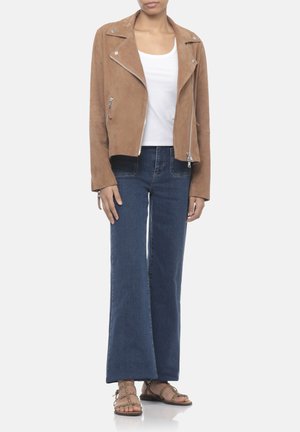 Persona con giacca moto in pelle scamosciata beige, canotta bianca, jeans blu a gamba larga e sandali marroni borchiati, in piedi davanti a uno sfondo semplice.