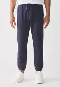 Man wearing navy blue jogger pants with elastic cuffs and white sneakers, standing against a plain white background.