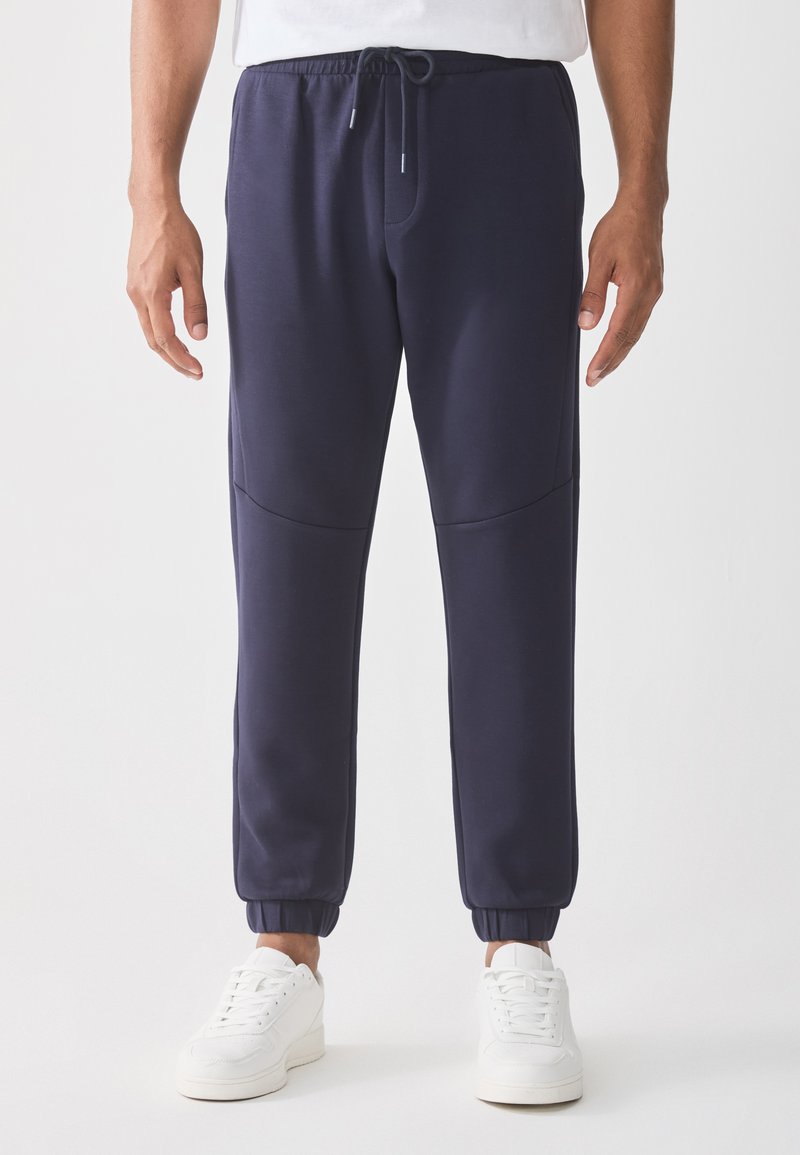 Man wearing navy blue jogger pants with elastic cuffs and white sneakers, standing against a plain white background.