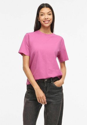 Jeune femme souriante, portant un t-shirt rose uni à manches courtes et un jean foncé, avec une main dans la poche.