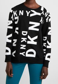 Zwart sweatshirt met verspreide grote witte DKNY-letters, gedragen met teal gekleurde broek, waarbij de romp en armen van een persoon zichtbaar zijn.