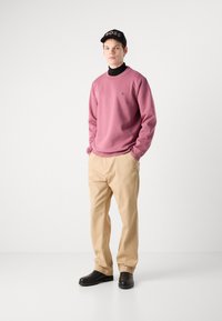 Felpa rosa chiaro con polsini a coste, collo alto nero, pantaloni beige e scarpe nere. Il cappellino è nero con il logo "BOSS" in rilievo.