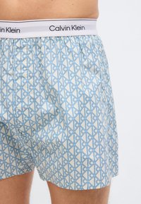 Κοντινό πλάνο ενός ατόμου που φορά ανοιχτόχρωμα μποξέρ Calvin Klein με μπλε γεωμετρικό μοτίβο και λευκή μέση με το όνομα της μάρκας.