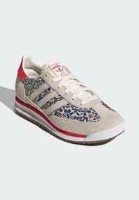 Beige mockasinsneaker med blommönstrade textilpaneler, röda detaljer, vita snören och en gummisula. Har tre signaturränder.