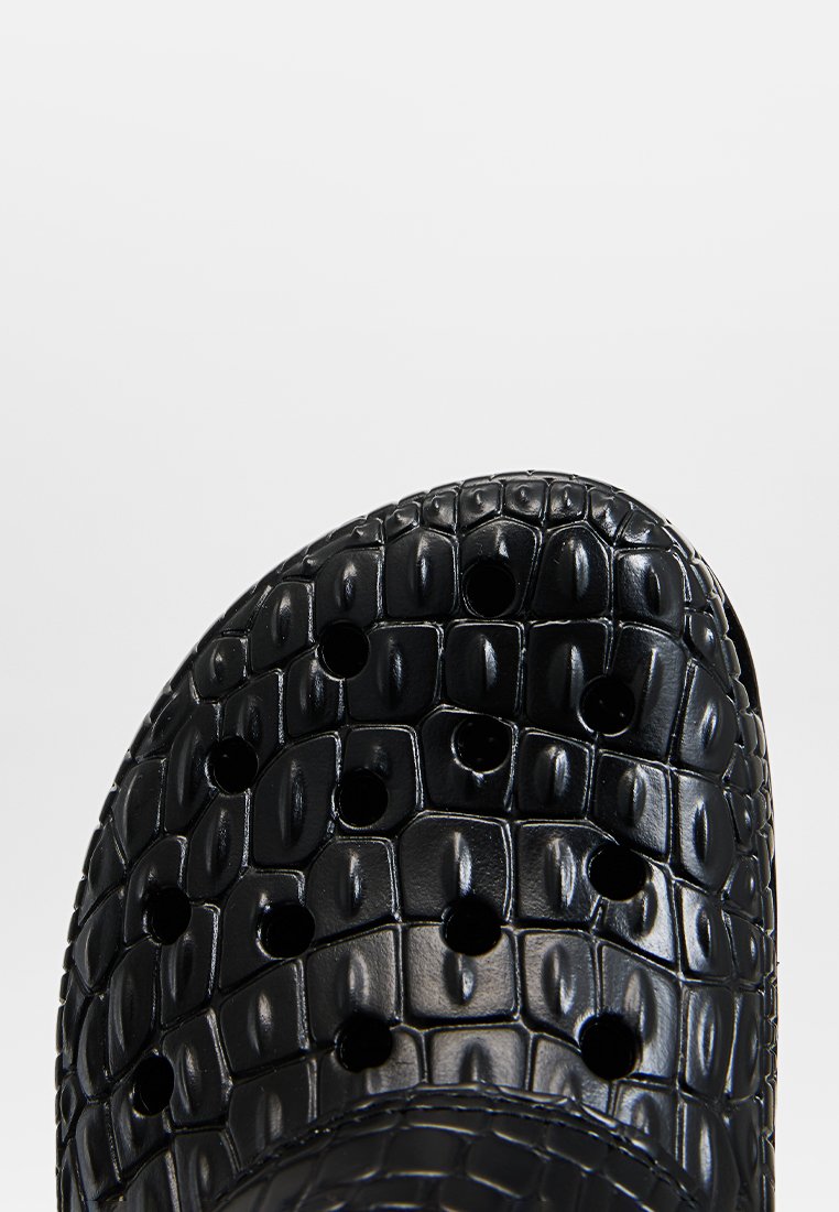 Zuecos negros hechos de un material texturizado con un patrón de cocodrilo, que cuentan con múltiples orificios de ventilación y un diseño de punta redondeada.