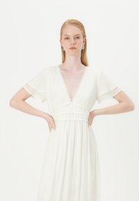 Vestido branco com mangas curtas e evasê, e um decote em V profundo, apresentando uma cintura franzida e detalhe de renda ao longo do decote. Textura de tecido suave.