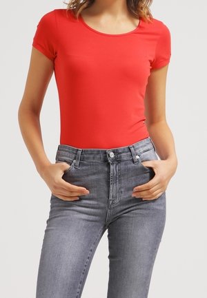 Kobieta w dopasowanym czerwonym t-shircie z krótkim rękawem i szarych wysokich jeansach, z rękami w przednich kieszeniach, stojąca na jednolitym tle.