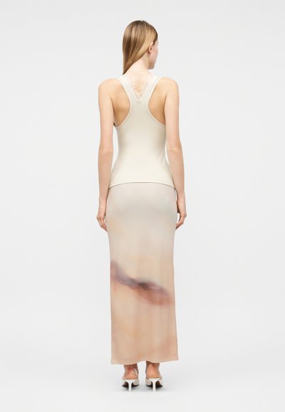 Γυναίκα που φαίνεται από πίσω, φορώντας ένα αμάνικο μπεζ τοπ με δαντελένιες λεπτομέρειες και μια μακριά φούστα με διαβαθμισμένο χρώμα, συνδυασμένη με μπεζ τακούνια.